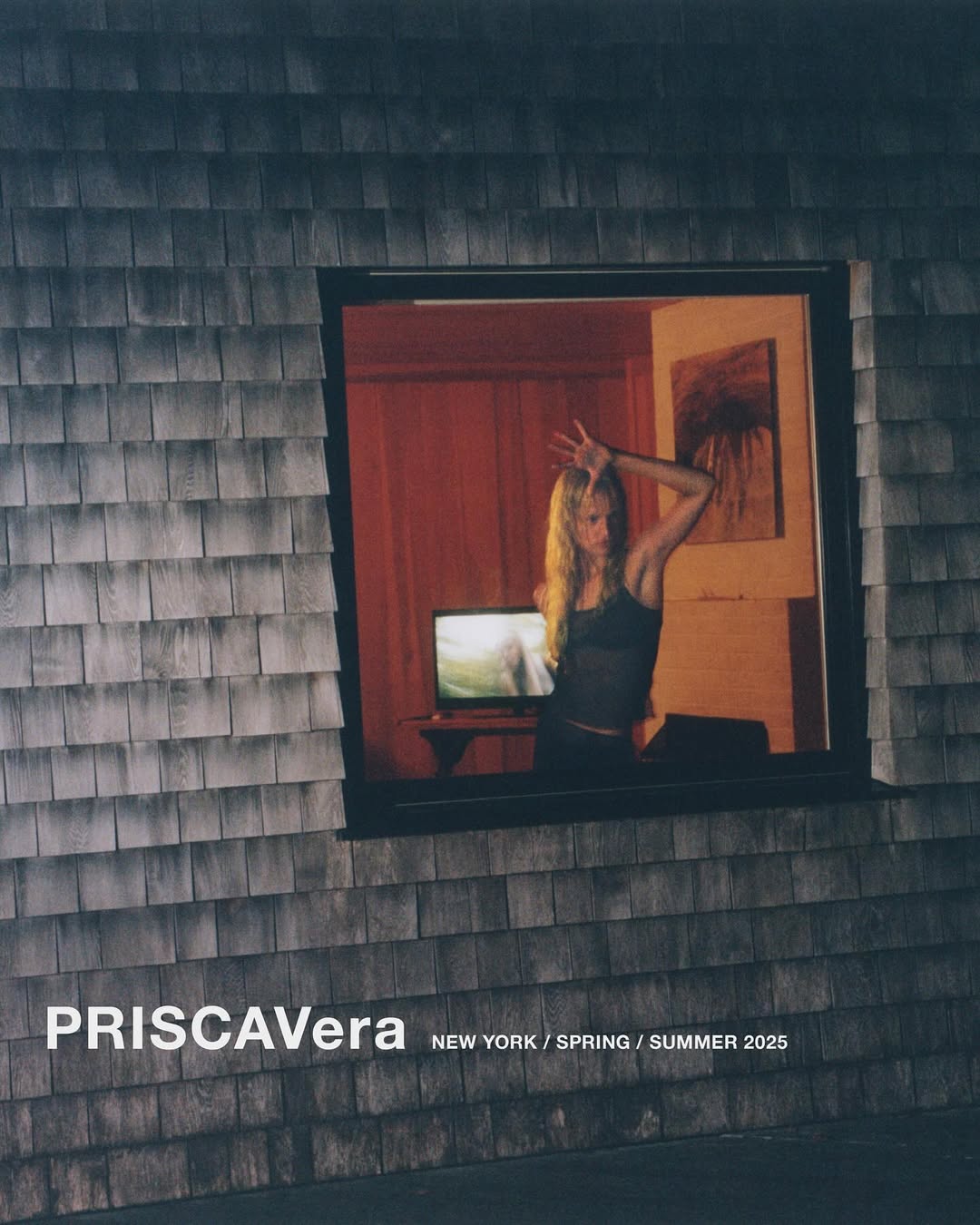 A woman poses behind a window in a gray shingled building with a television on a table inside a warmly lit, red-walled room.