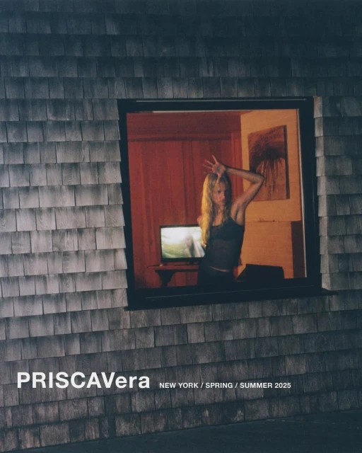 A woman poses behind a window in a gray shingled building with a television on a table inside a warmly lit, red-walled room.
