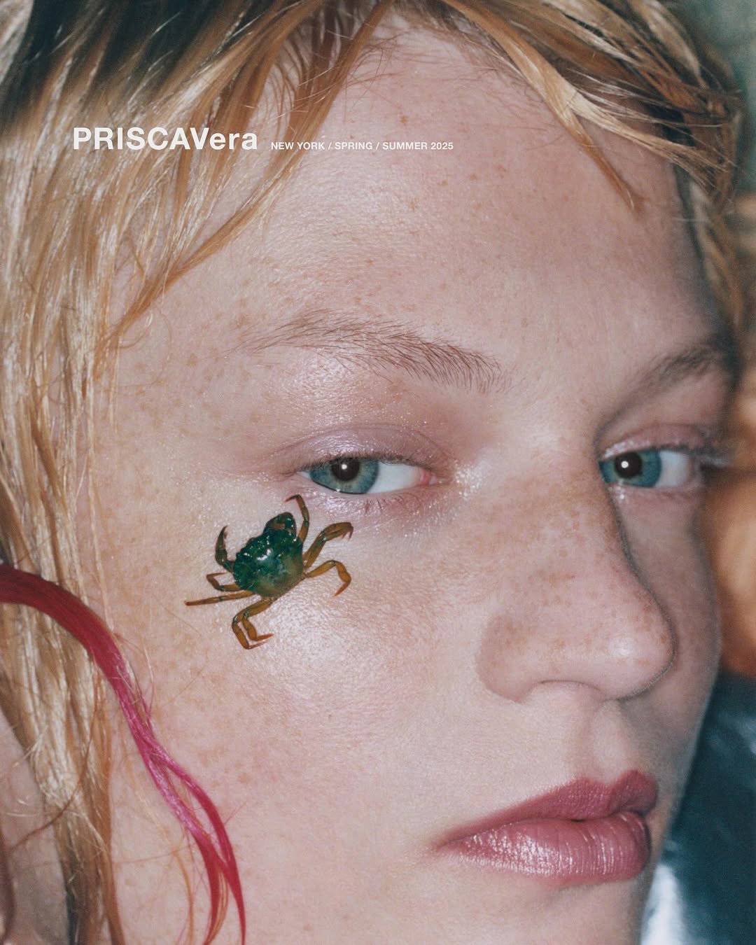 Close-up of fair-skinned female model with a small green crab affixed to her cheek, gazing forward with pink hair accents.