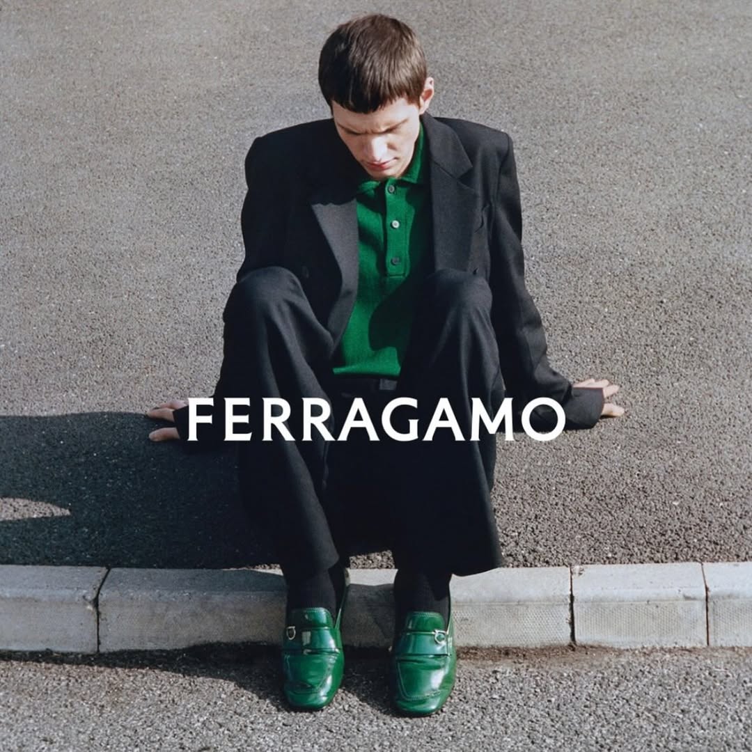Fashion editorial shot of a young adult man in a black overcoat and green polo shirt sitting on a concrete curb in an urban setting.