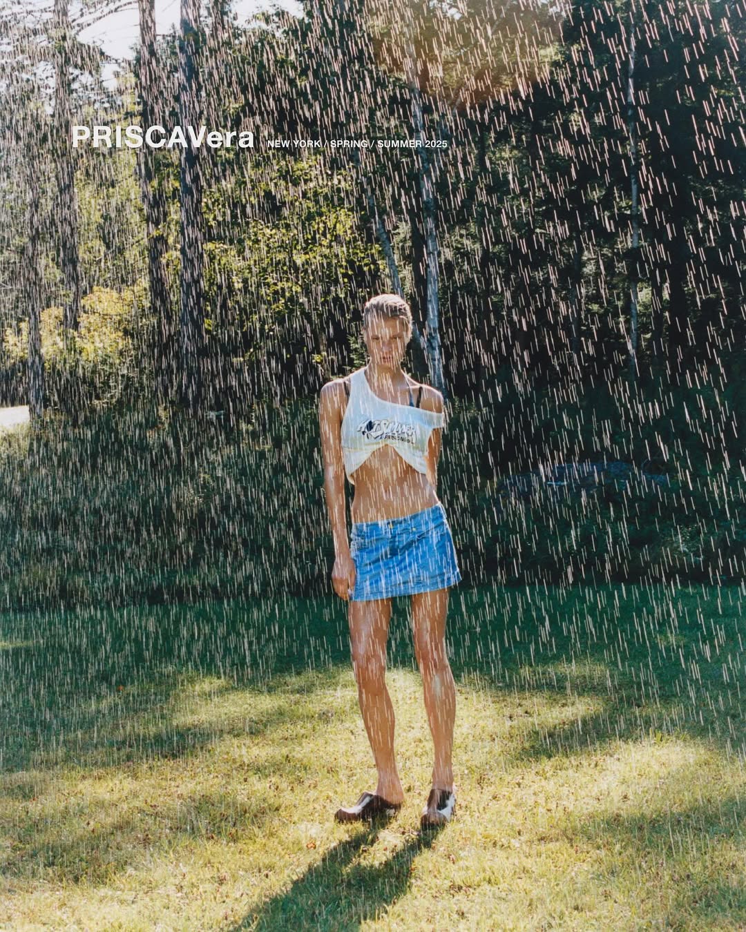 A woman stands in a green field under artificial rain wearing a white top and jean skirt, surrounded by trees in the background.