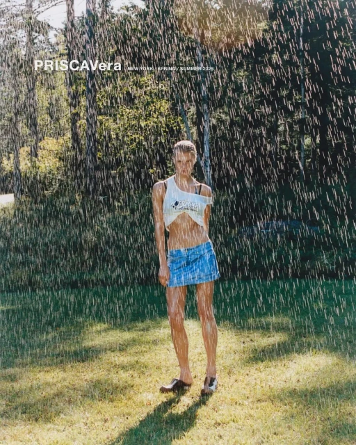 A woman stands in a green field under artificial rain wearing a white top and jean skirt, surrounded by trees in the background.