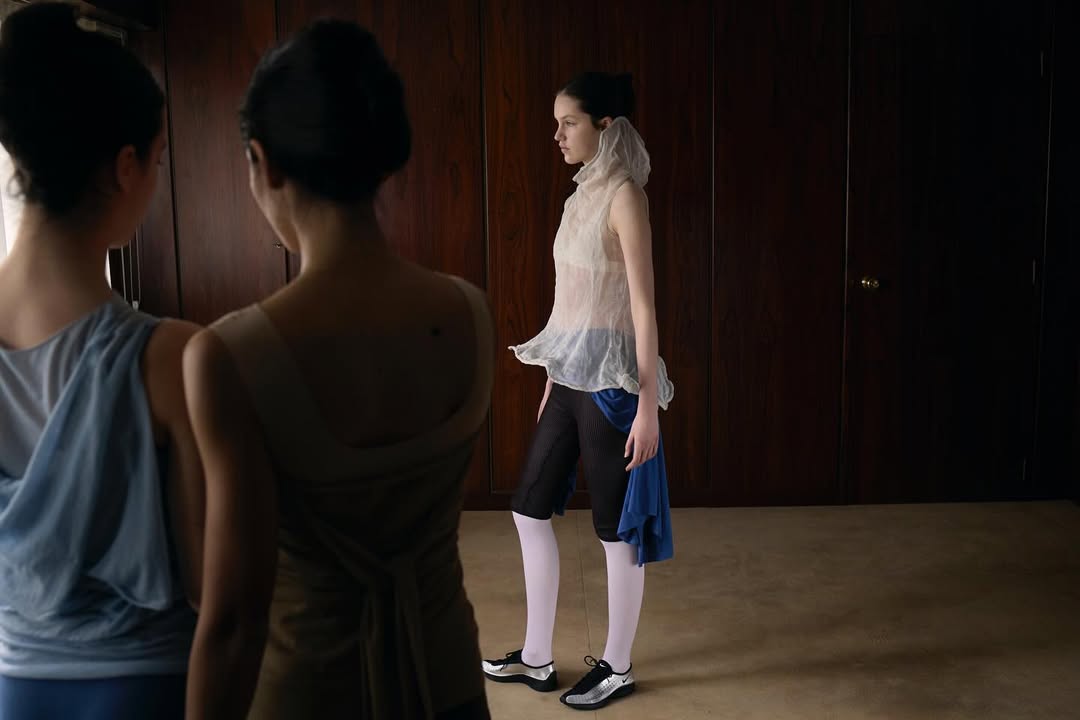 Three women in avant-garde fashion indoors against wood paneled walls and carpeted floor.