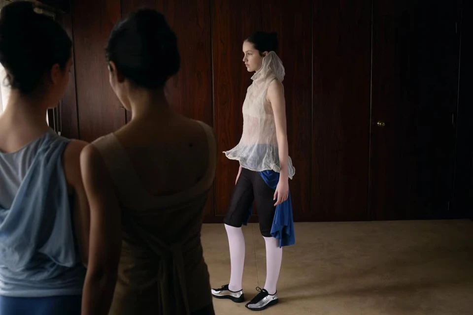 Three women in avant-garde fashion indoors against wood paneled walls and carpeted floor.