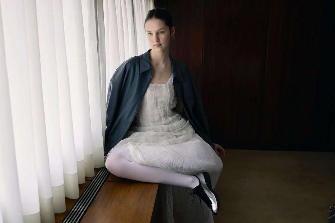 Young woman wearing a dark blue leather jacket and white dress seated near a white curtained window by Julia Asaro