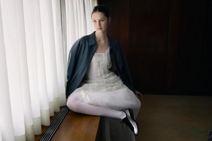 Young woman wearing a dark blue leather jacket and white dress seated near a white curtained window by Julia Asaro