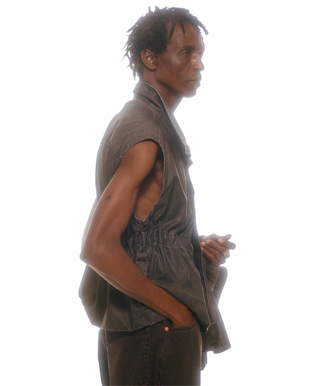 Man in sleeveless shirt and dark pants in a side view against a white background. Fashion-editorial style with neutral lighting.