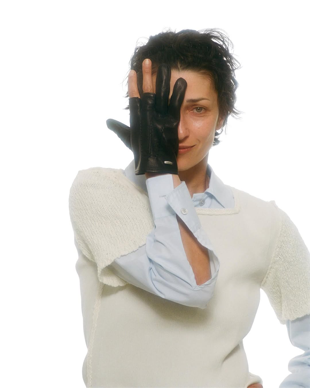 Woman with short dark hair covers half her face with a black leather, fingerless glove, wearing a white sweater and light blue collared shirt.