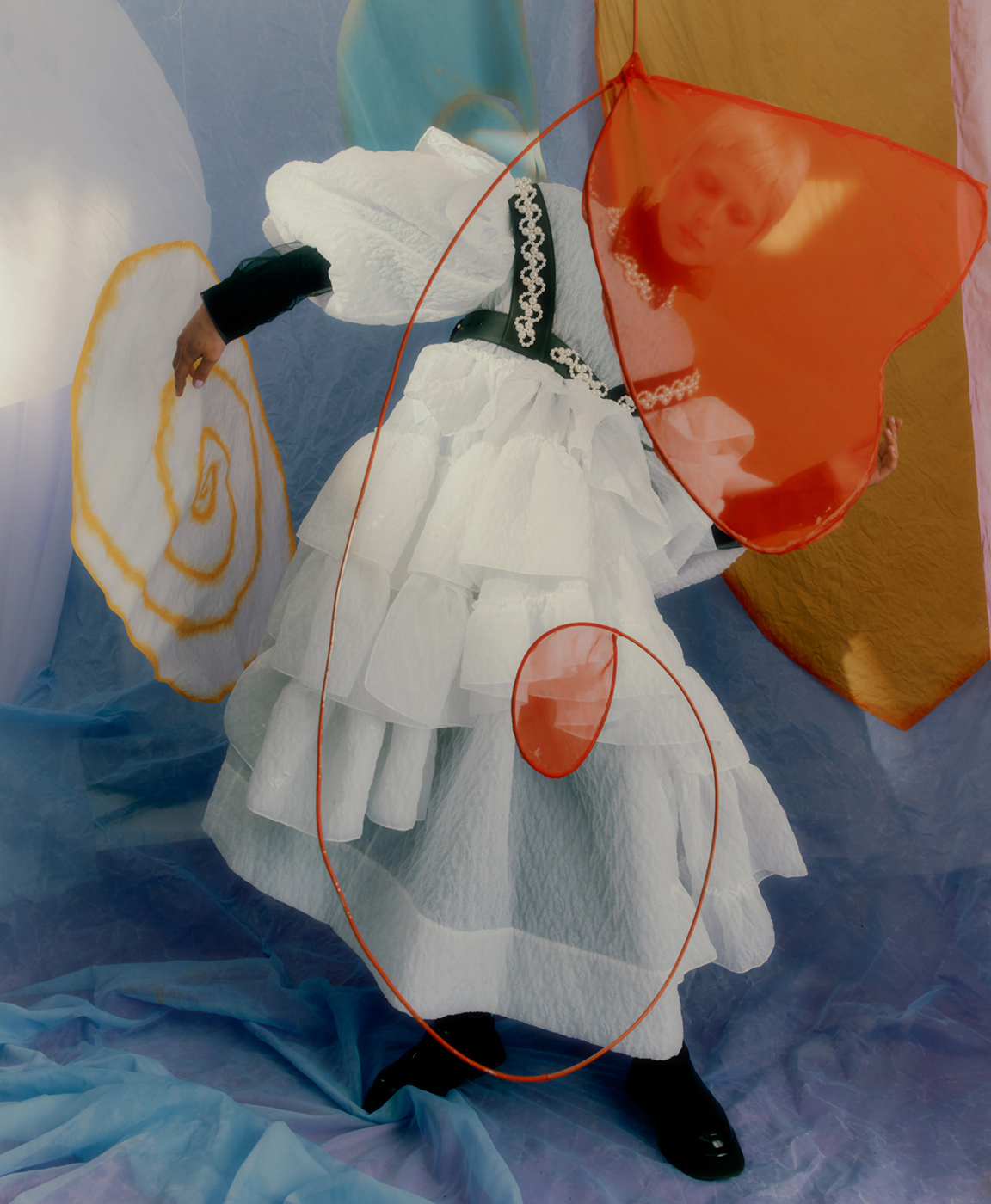 Fashionable person in white ruffled dress with black accents posing with orange translucent heart and white spiral prop in studio with blue backdrop.