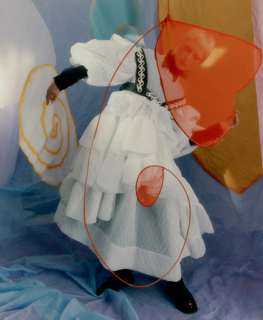 Fashionable person in white ruffled dress with black accents posing with orange translucent heart and white spiral prop in studio with blue backdrop.