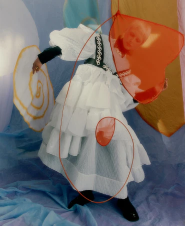 Fashionable person in white ruffled dress with black accents posing with orange translucent heart and white spiral prop in studio with blue backdrop.
