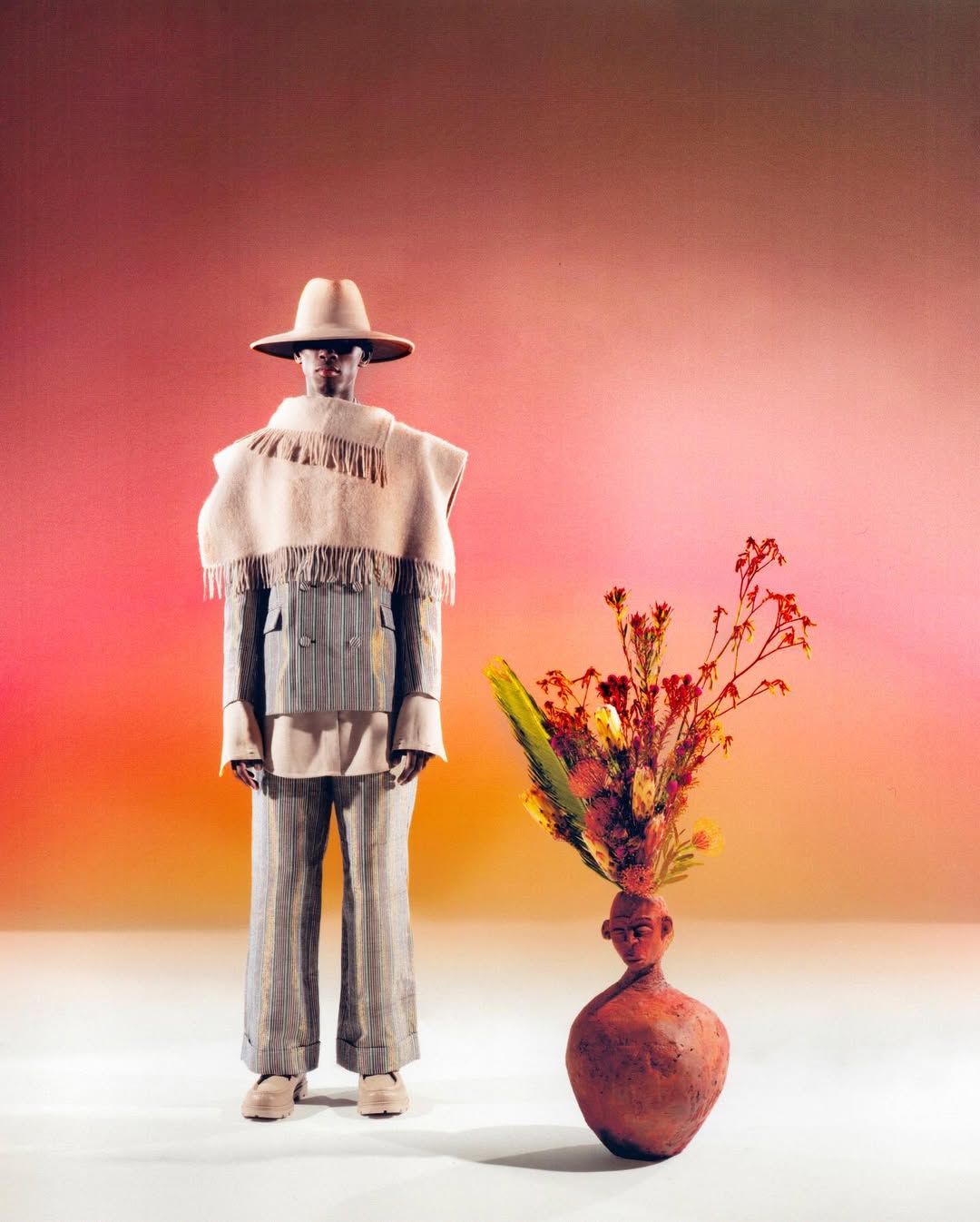 Man in a patterned suit and hat stands next to a vase of flowers in front of a mottled pink background in a studio fashion shot.