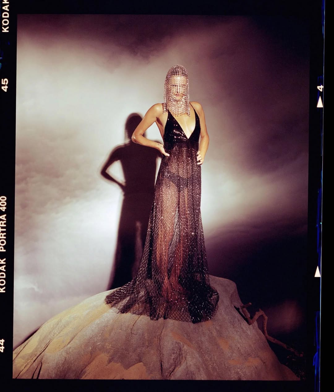 Elegant woman in sheer sequin gown poses on a rock formation in a photo studio setting with a dramatic shadow backdrop.
