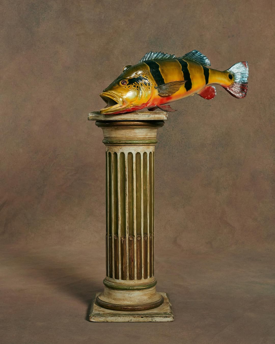 A colorful fish sculpture stands on a weathered, fluted column against a mottled brown backdrop, creating a still life with artistic appeal.