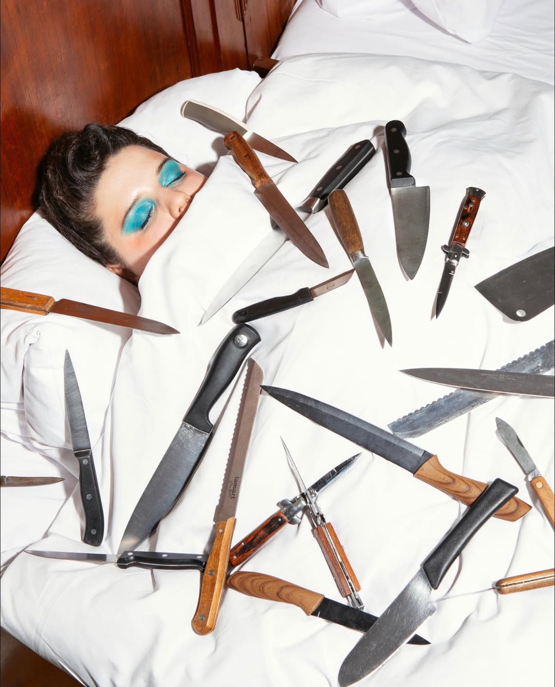 Person sleeping in a bed surrounded by knives with blue eye makeup, white bedding, and a wooden headboard in a brightly lit room.