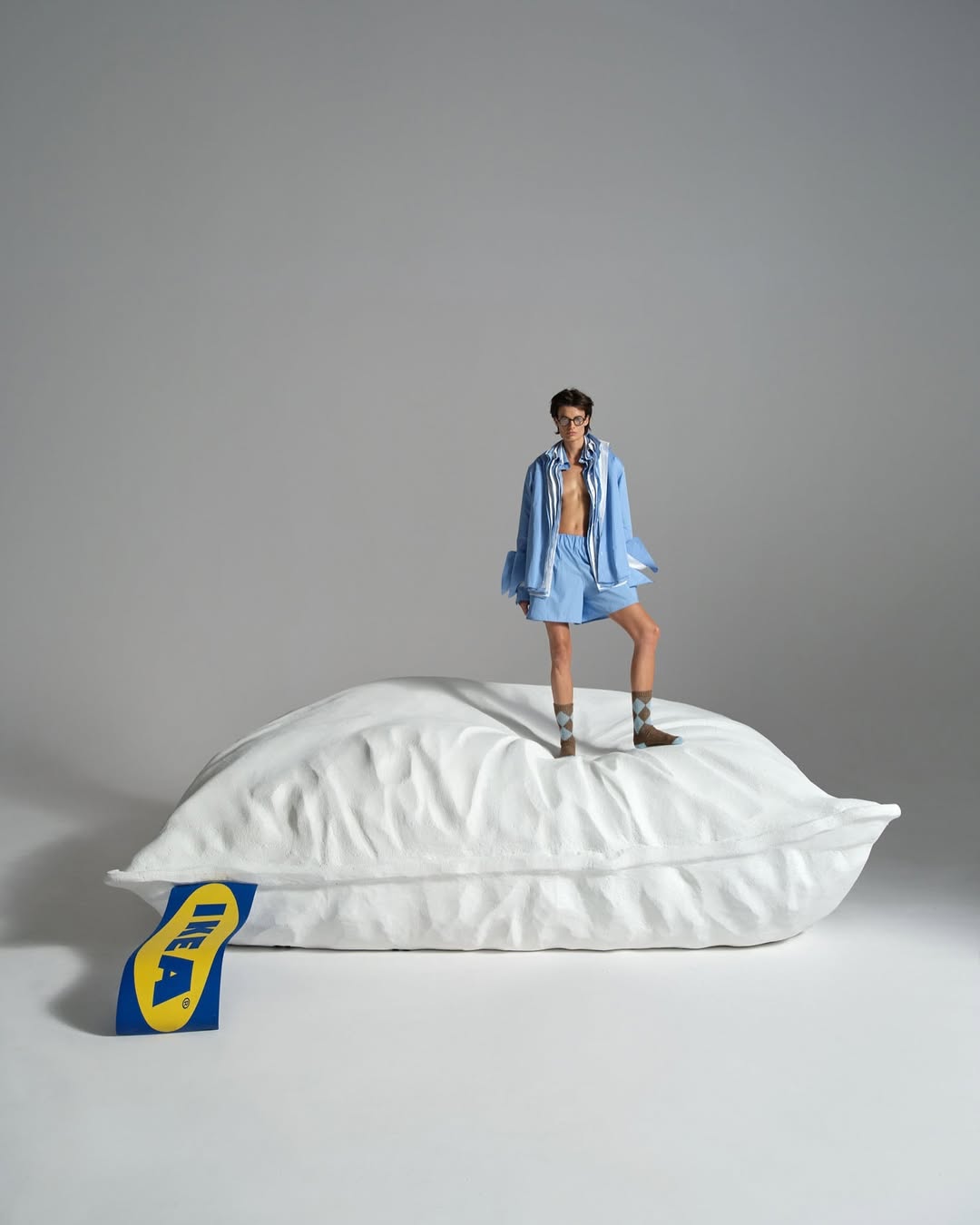 Adult male poses on a white pillow with an IKEA logo tag in a studio setting wearing a light blue short set, brown socks, and dark glasses.