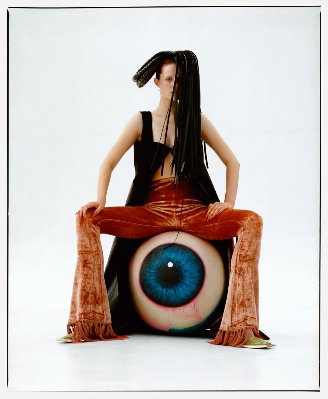 Fashion photo of woman with unique hairstyle wearing black top, brown pants, and sitting on giant eyeball in a white studio.