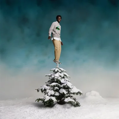 A smiling black man standing atop a small snow-covered pine tree against a textured teal gradient background.
