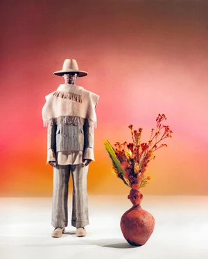 Model wearing a wide-brim hat, fringed poncho, and striped suit standing beside a clay vase holding red flowers