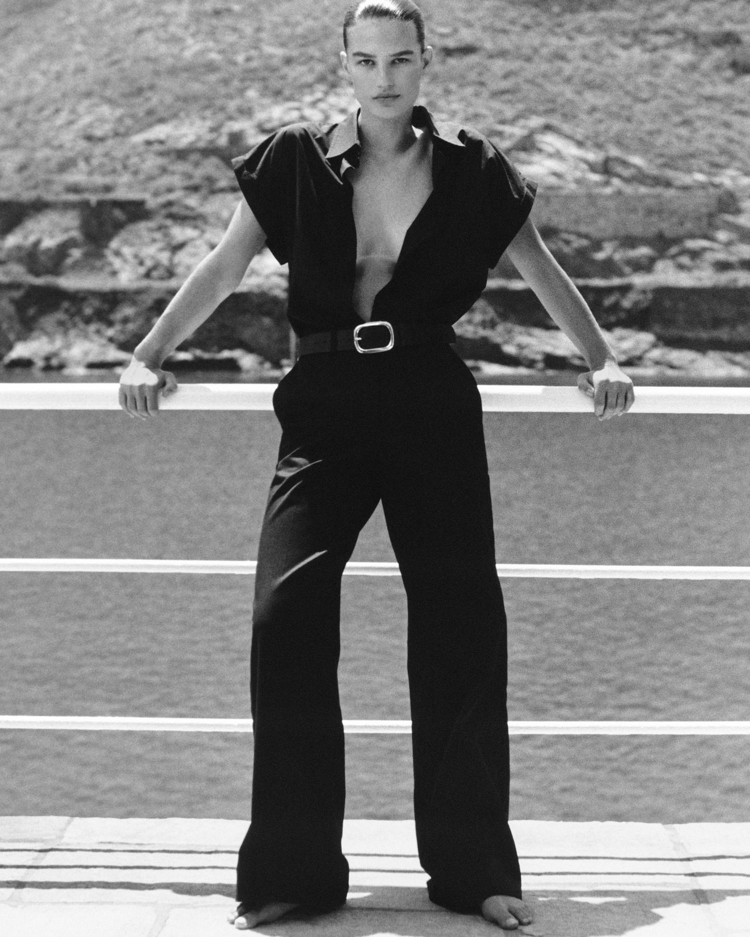 A fashionable woman stands on a balcony in a black outfit and belt, with a staircase in the background, captured in black and white.