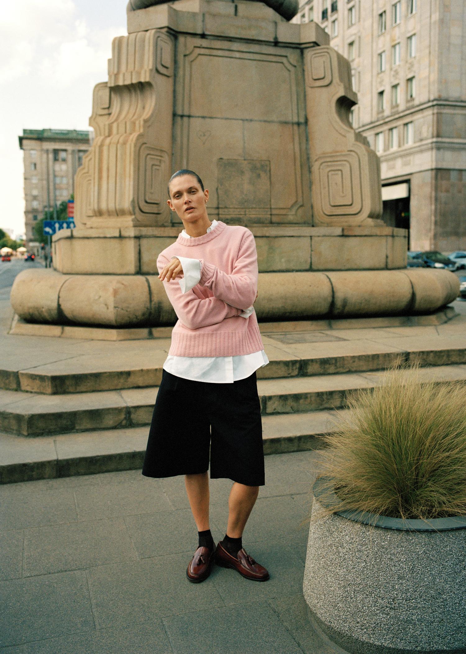 Model wearing a pink sweater and black shorts stands in front of a large stone structure in a city street setting.