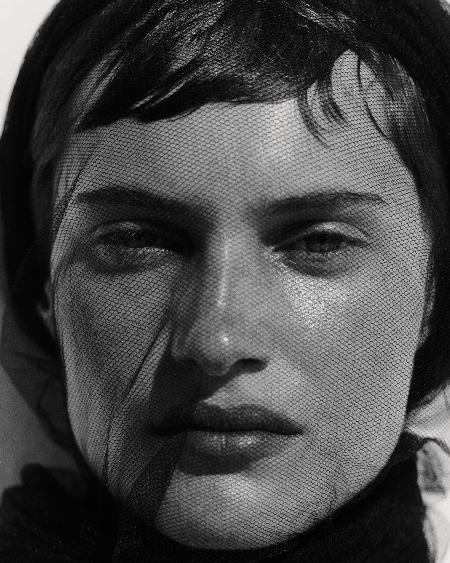 Close-up black and white portrait of a young woman with black netting over her face in a studio with neutral lighting.