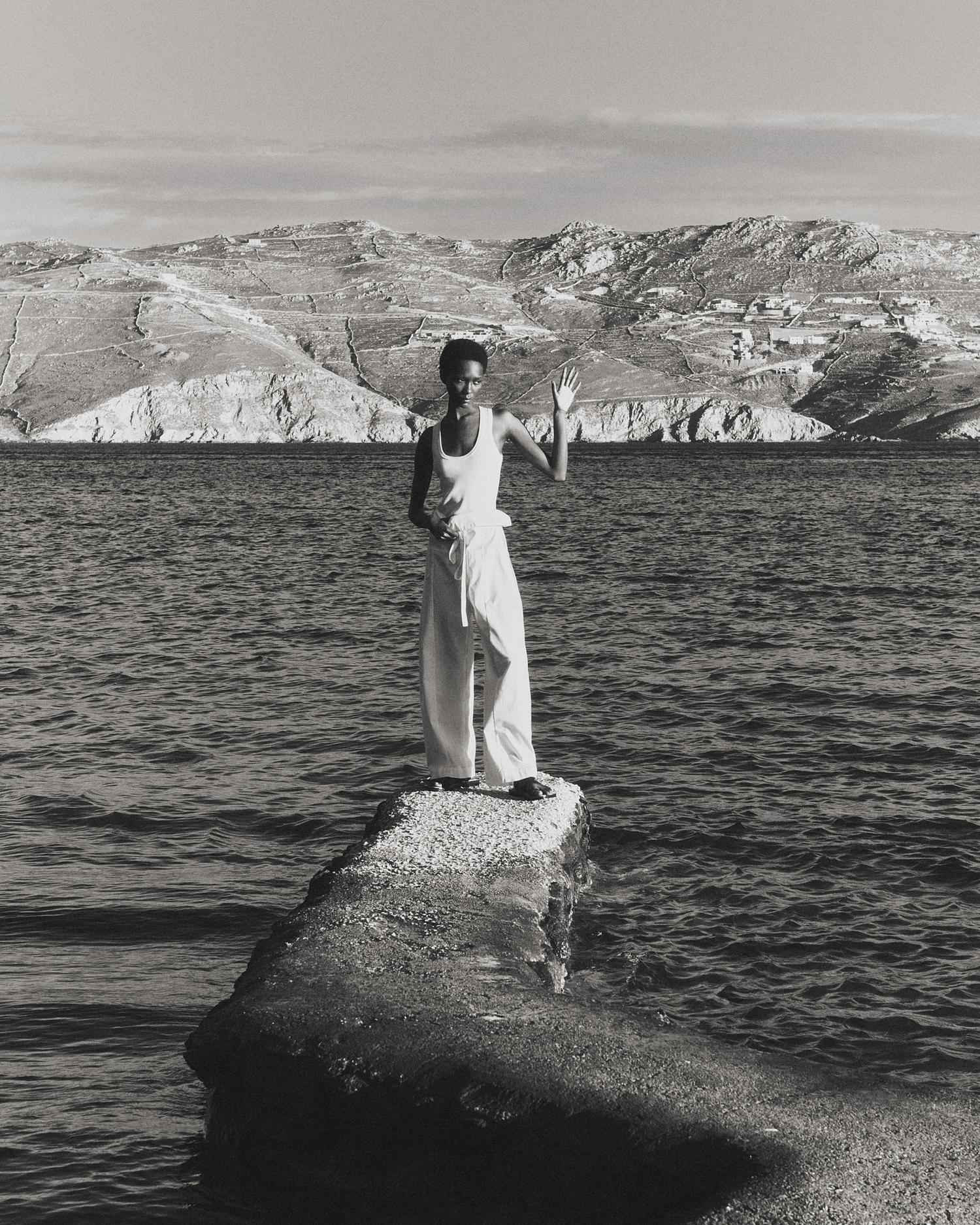 Monochrome photograph of a Black woman standing on a concrete pier with her arm raised in front of a sea and mountainous landscape.