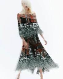 Model with long blonde hair and dark lipstick wearing a patterned off-the-shoulder knitted dress against a bright white background.