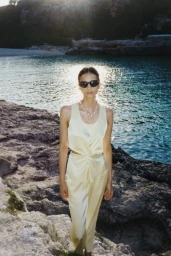Woman wearing sunglasses and a pale yellow top standing by rocky shoreline at sunset