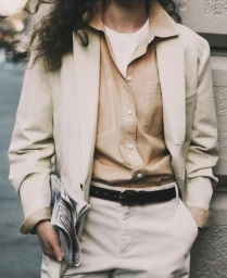 Person wearing a cream blazer, tan button-down shirt, and white t-shirt holding folded newspapers outside.