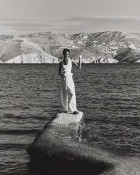 Black woman in white tank top and trousers standing on a concrete pier by the water with distant hills.