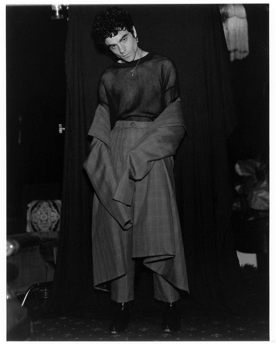 Monochrome portrait of a male model in a sheer black shirt, oversized plaid trousers, and a draped blazer against a dark backdrop.