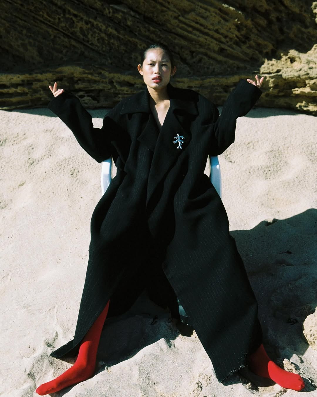 Asian female model in black coat and red stockings poses on beach in natural light, sitting in a chair with rocky background.