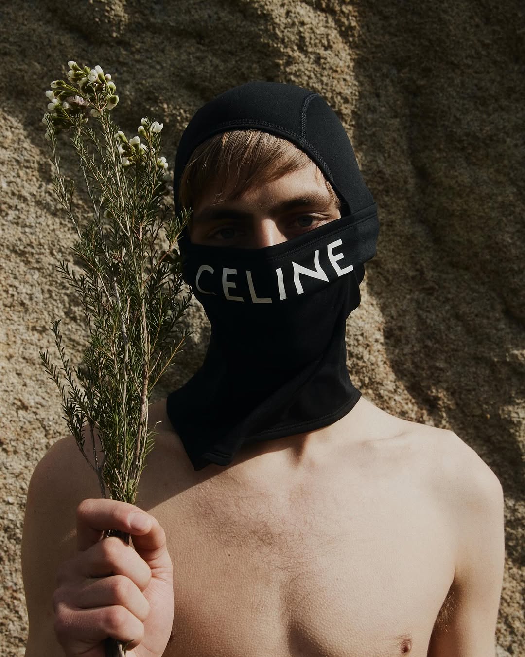 Nude male fashion model in black balaclava with white 'CELINE' text holds flowers against a textured rock background in an outdoor editorial photoshoot.