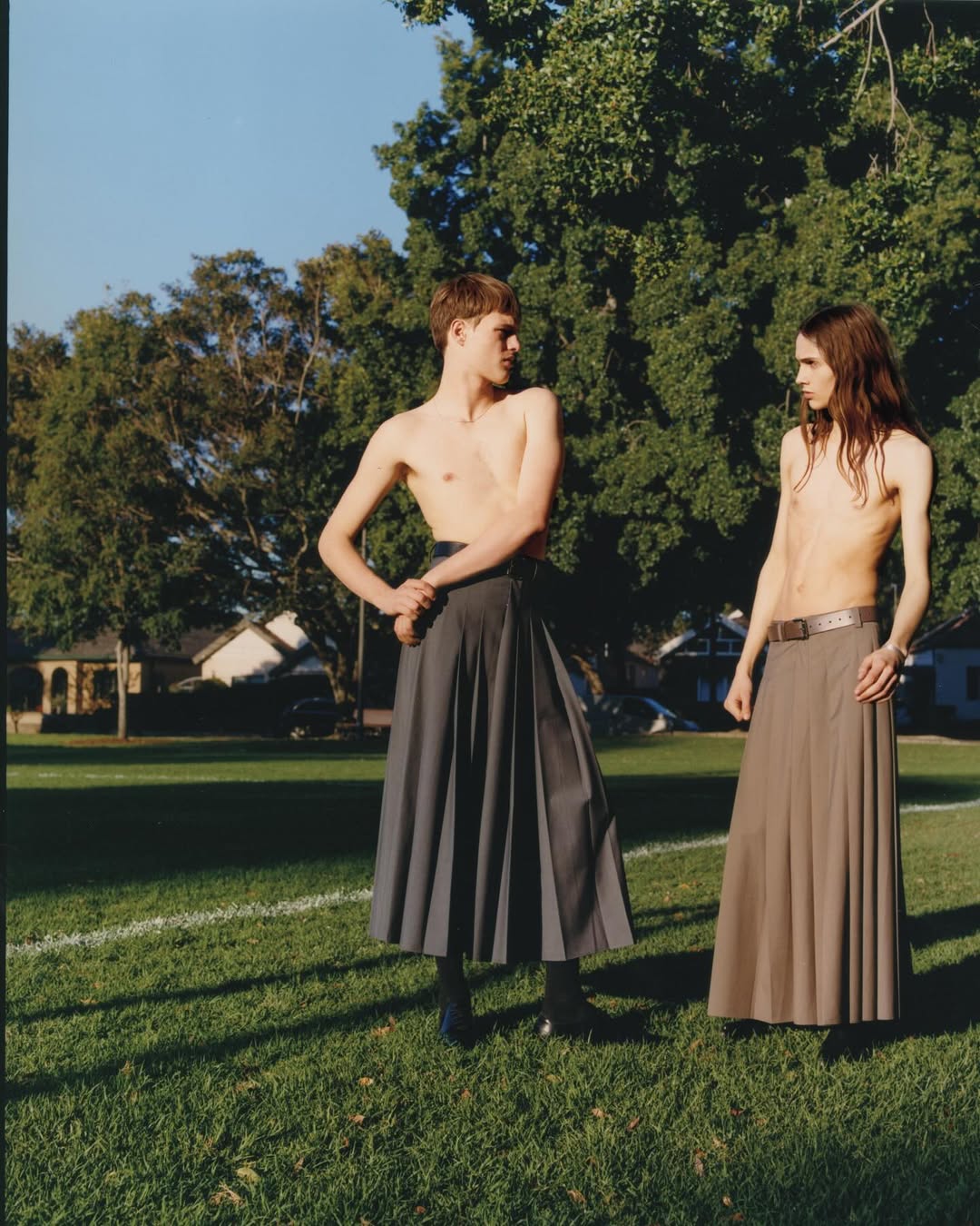 Two shirtless young men in skirts stand on a grassy field with a tree and houses in the background, captured in an outdoor setting.