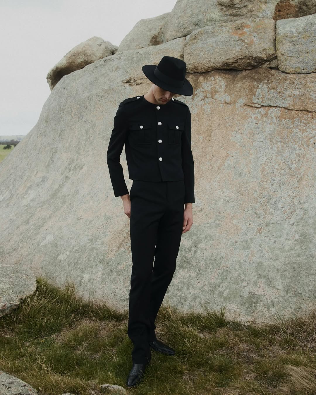 A tall, slender man in a black suit and hat poses outdoors on a grassy field with a large boulder, creating a striking fashion editorial.