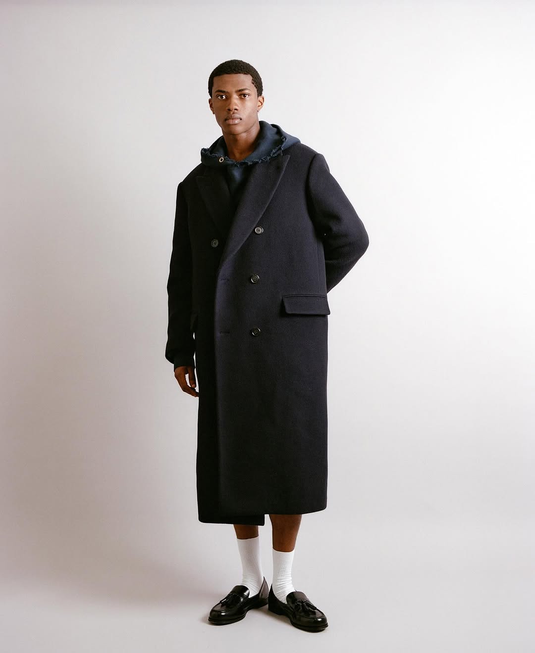 Studio shot of a Black male model wearing a long navy coat, navy hoodie, white socks, and black loafers posing against a plain background.