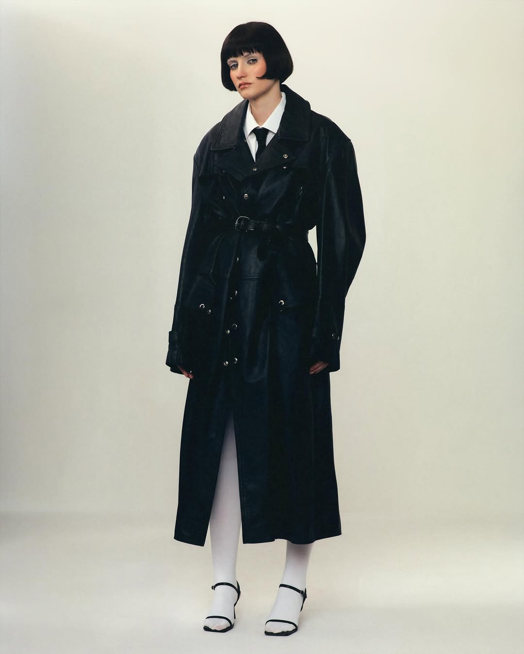 A fashionable woman with a bob haircut poses in a belted black trench coat, white shirt and tie, and white tights with black heeled sandals.