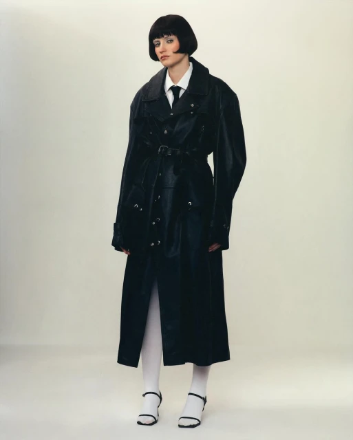 A fashionable woman with a bob haircut poses in a belted black trench coat, white shirt and tie, and white tights with black heeled sandals.