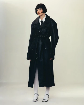 A fashionable woman with a bob haircut poses in a belted black trench coat, white shirt and tie, and white tights with black heeled sandals.