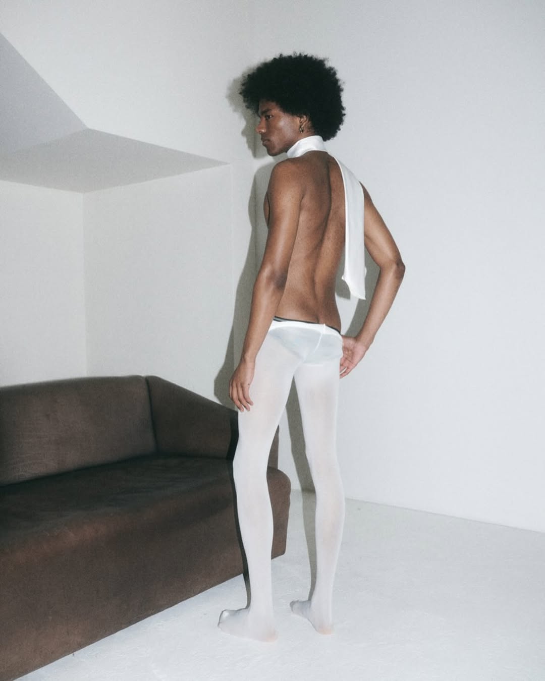 A slender African-American man with an afro poses in white tights with a towel draped around his neck. He stands near a brown couch.