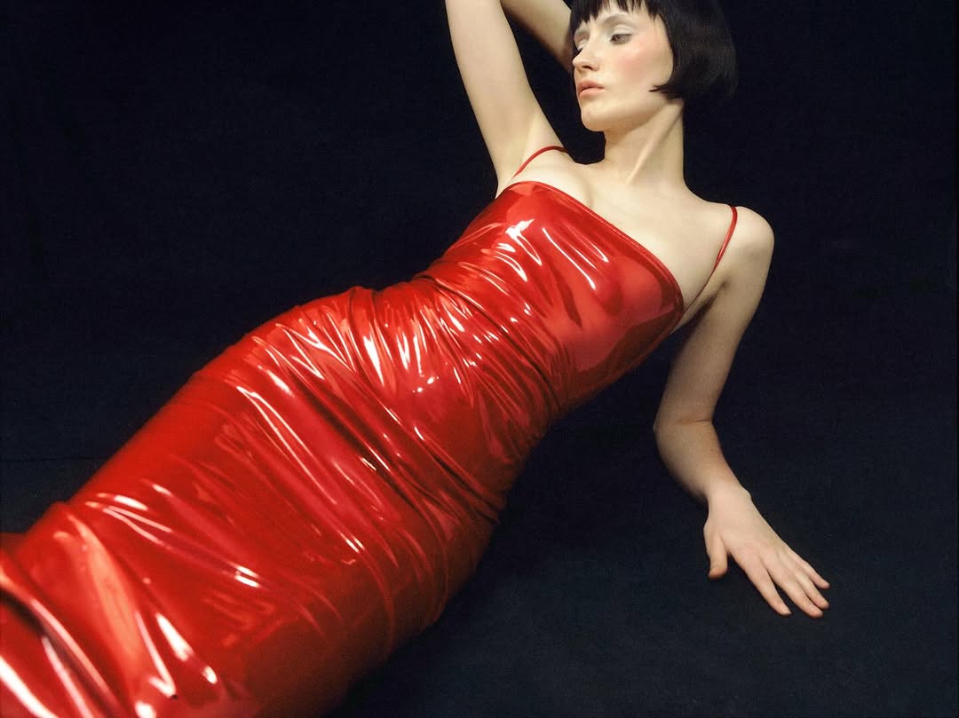 A slender woman in a red, shiny dress lies against a black backdrop, her arm raised, looking off-camera in a fashion or beauty editorial.