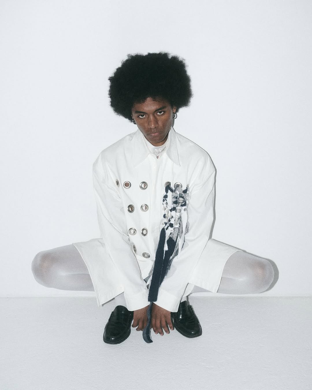 Person with an afro squats in front of a white background wearing a white button-down shirt, white pants, and black patent leather shoes.