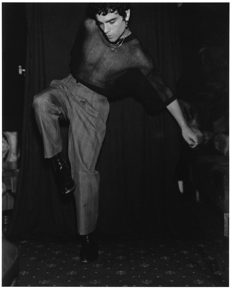 Monochrome image of a stylish adult male in motion, wearing a sheer top and patterned pants, set against a dark backdrop.