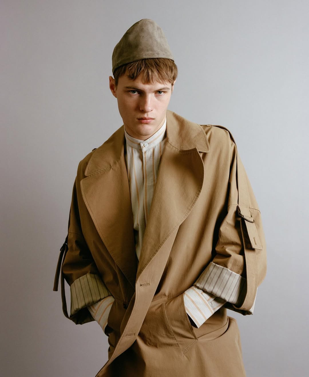 Male fashion model poses in a studio wearing a light brown trench coat, striped shirt, and matching hat against a neutral backdrop.