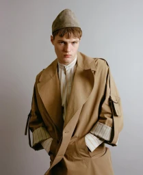 Model in camel trench coat and matching cap styled by Rosalind Seehusen against a plain grey studio backdrop