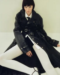 Model with dark bob wearing a black leather trench coat, white shirt, and white tights seated on a pale floor