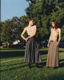 Two shirtless male models wearing pleated skirts standing on a green park lawn outdoors in Melbourne.