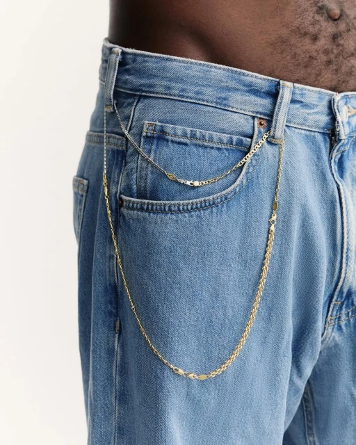Close-up of a person wearing light blue jeans with gold and silver metal chains attached to the belt loops and pocket.