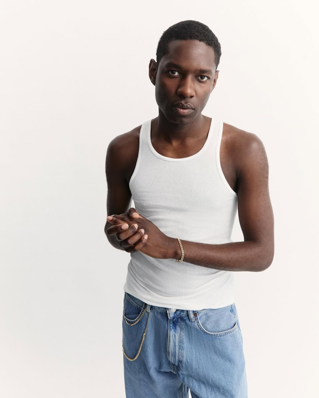 Portrait of a young Black male in a white ribbed tank top and light wash jeans, posing against a bright white backdrop in a studio setting.